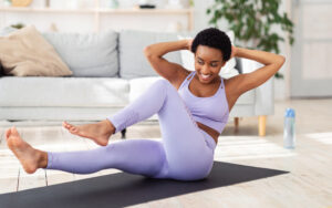 A woman in a purple workout outfit smiling while performing a bicycle crunch exercise on a yoga mat in a bright, comfortable living room.